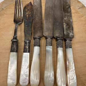 May include: A set of six vintage utensils including one fork and five knives with tarnished metal blades and white, fluted mother-of-pearl handles. Each piece features a decorative silver-toned metal collar with intricate etched detailing.