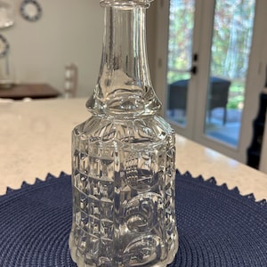 May include: A clear, decorative glass bottle with a detailed, geometric pattern. The bottle has a wide base, a rectangular body, and a long, slender neck. The top of the bottle has a rounded stopper. The bottle is sitting on a blue placemat.