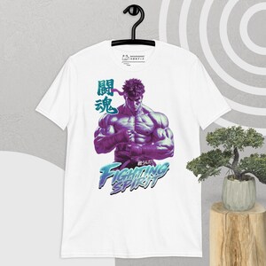 Op de afbeelding: Wit T-shirt met een afbeelding van een gespierd personage in paarse en blauwe tinten, Japanse tekst en de woorden "Fighting Spirit". Het ontwerp is gedrukt op een T-shirt met korte mouwen dat aan een zwarte hanger hangt.