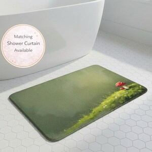 Peut inclure: Un tapis de bain rectangulaire avec un fond vert et un motif peint d'une zone herbeuse avec deux champignons à chapeau rouge. Le tapis est posé sur un sol carrelé blanc. Le texte sur l'image dit "Rideau de douche assorti disponible."