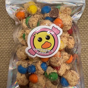 May include: A clear, iridescent bag filled with candy. The candy is a mix of round, textured pieces and colorful, coated candies in shades of blue, orange, yellow, green, and red. A sticker with a duck logo and the text "QUACKER'S CANDY" is on the bag.