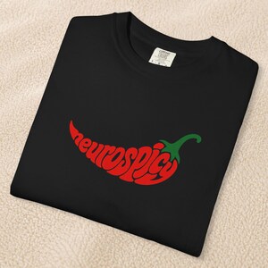 May include: A black t-shirt featuring a graphic of a red chili pepper shaped by the text "neurospicy" in a bold, lowercase font. The stem of the chili pepper is green. The shirt has a white interior neck label with visible text and size L.