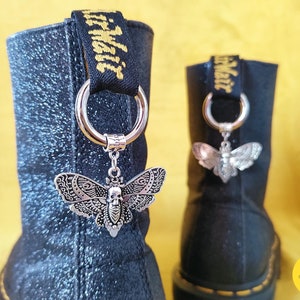 Könnte beinhalten: Schwarze Lederstiefel mit silbernen Schmetterlingsanhängern an den Schnürsenkeln. Die Anhänger haben ein Totenkopfdesign und komplizierte Details. Die Stiefel haben eine schwarze Glitzer-Oberfläche und der Text "Happy Lost" ist auf dem rechten Stiefel sichtbar.