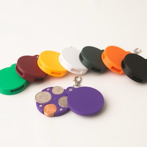 May include: A set of eight colourful coin holders with a keychain. The holders are in various colours, including green, brown, yellow, white, black, and orange. One holder is open, revealing a compartment for coins.