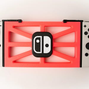 May include: A red and black protective case for a gaming console. The case has a geometric design and a black logo in the center. The console's white controllers are visible on either side.