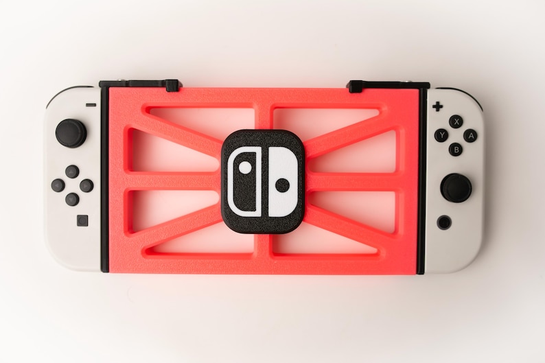 May include: A red and black protective case for a gaming console. The case has a geometric design and a black logo in the center. The console's white controllers are visible on either side.