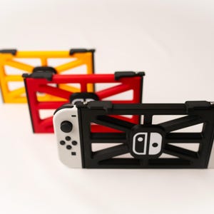 May include: Three game console holders in yellow, red, and black. The black holder is attached to a white and black game console. The holders have a geometric design and the game console has a white logo.