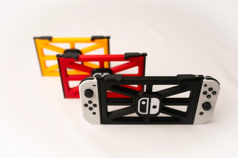 May include: Three game console holders in yellow, red, and black. The black holder is attached to a white and black game console. The holders have a geometric design and the game console has a white logo.