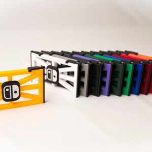 May include: A collection of colorful Switch console stands. The stands are in various colors, including yellow, white, gray, black, purple, green, blue, red, pink, and orange. Each stand features a Switch logo.