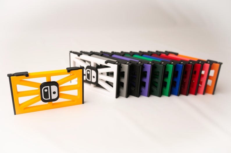 May include: A collection of colorful Switch console stands. The stands are in various colors, including yellow, white, gray, black, purple, green, blue, red, pink, and orange. Each stand features a Switch logo.