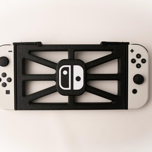 May include: A black and white Nintendo Switch console with a black frame. The frame has a geometric design and surrounds the console's central logo. The controllers are white with black buttons.