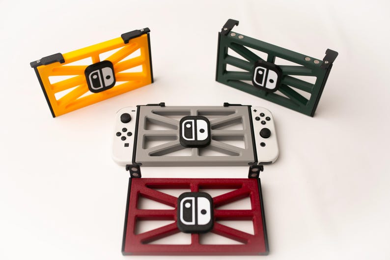 May include: A collection of colorful Nintendo Switch accessories. The image features a yellow, green, and red frame-like holder, each with a black and white logo. A gray holder is attached to a white Switch console.