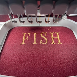 Puede incluir: Una m&aacute;quina de bordar en funcionamiento, bordando la palabra "FISH" en hilo dorado sobre una tela burdeos. Las agujas y el marco de la m&aacute;quina son visibles, destacando el proceso detallado de creaci&oacute;n de texto personalizado.
