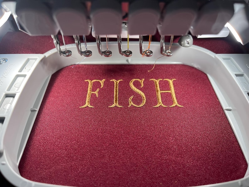 Puede incluir: Una m&aacute;quina de bordar en funcionamiento, bordando la palabra "FISH" en hilo dorado sobre una tela burdeos. Las agujas y el marco de la m&aacute;quina son visibles, destacando el proceso detallado de creaci&oacute;n de texto personalizado.