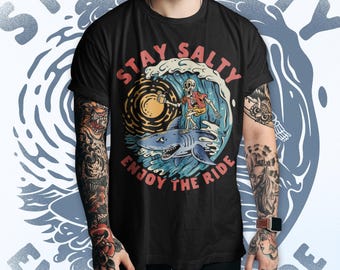 May include: Black t-shirt with a graphic featuring a skeleton surfer riding a shark on a wave. The design includes the text "Stay Salty Enjoy the Ride" in red and white.