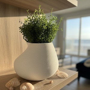 May include: A white, textured vase with a spiral design, filled with green plants, sits on a wooden shelf. Seashells and sand are scattered around the vase. The background shows a bright, sunny room with a view of the ocean.