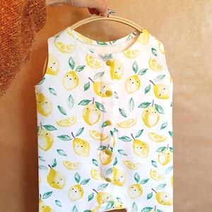 May include: White sleeveless romper with a lemon pattern. The fabric features yellow lemons with smiling faces, green leaves, and lemon slices. The romper has yellow buttons down the front and is held by a wooden embroidery hoop.