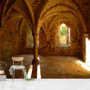 May include: Interior view of a stone-walled room with arched ceilings and pillars. Sunlight streams through an arched doorway and window, lighting the room. A wooden chair with a green cloth draped over it is in the foreground.