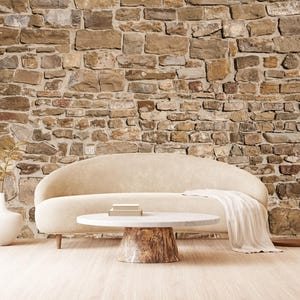 Op de afbeelding: Een lichtbeige gebogen bank met een witte deken staat voor een stenen muur. Een ronde, witte salontafel met een houten onderstel staat voor de bank.