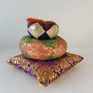 May include: Three decorative cushions stacked on top of each other. The bottom cushion is square, purple with gold floral patterns. The middle cushion is round, with green, orange, and gold floral patterns. The top cushion is small, round, navy blue with gold accents.