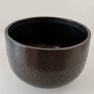 May include: A dark brown bowl with a textured exterior and a smooth, black interior. The bowl has a rounded shape and a wide opening. The exterior has a dimpled pattern. The bowl is likely made of metal or a similar material.