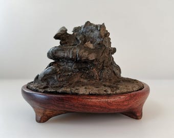 May include: A unique bonsai sculpture featuring a dark, textured, organic form atop a rich, reddish-brown wooden base. The base has a rounded shape with three small feet, creating a natural, earthy aesthetic.