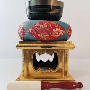 May include: A meditation bowl set. The black and gold bowl sits on a patterned cushion, placed on a gold stand. A wooden mallet with a red handle and white striking end rests below. The cushion has a red floral design.
