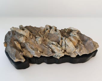 May include: A natural, irregularly shaped rock formation with a variety of colors, including gray, tan, and brown. The rock sits on a black, rectangular base. This decorative piece could be used for home decor or as a display.