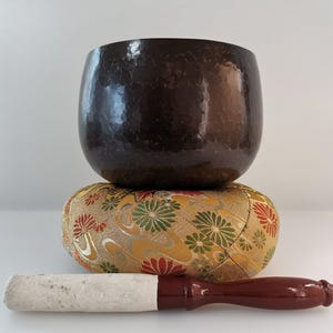 May include: A dark brown singing bowl with a hammered finish sits on a gold and green patterned cushion. A white wooden striker with a red handle is in front of the bowl.