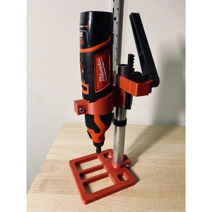 May include: A red and black drill press stand with a Milwaukee drill attached. The stand has a base with three slots and a vertical post with a clamp to hold the drill.