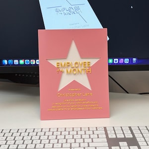 Puede incluir: Un premio rosa de "Empleado del Mes" con un recorte de estrella, con letras doradas y el nombre "Christopher Lane". El premio está sobre una superficie blanca, con un teclado en primer plano y un monitor de ordenador en el fondo.