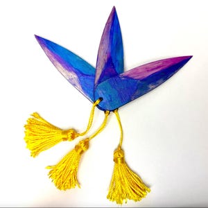 Puede incluir: Un marcapáginas artesanal abstracto con un diseño floral. El marcapáginas presenta tres elementos en forma de pétalo pintados en tonos azules y morados. Borlas amarillas brillantes cuelgan de la base. El marcapáginas está sobre un fondo blanco.