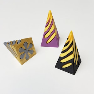 Pode incluir: Três peças de arte geométrica. Uma é um cubo com um desenho de flor, e as outras são em forma de pirâmide. As pirâmides são pretas, roxas e douradas com um padrão listrado. O cubo é dourado e cinza.