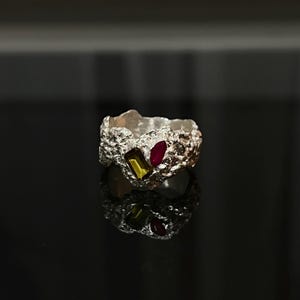 May include: A textured silver ring with a rough, organic design. The ring features two rectangular gemstones: one yellow-green and one red. The ring is displayed on a reflective black surface.