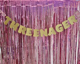 May include: Gold glitter letters spelling "THREE-NAGER" on a pink and white tinsel backdrop.