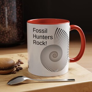 May include: A white ceramic mug with a bright red interior and handle. The exterior features the black text Fossil Hunters Rock! next to a large, intricate spiral graphic composed of numerous small black dots resembling a fossilised shell.
