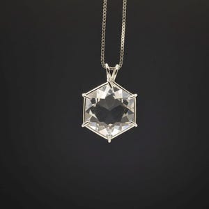 Op de afbeelding: Een heldere, facetgeslepen kristallen hanger in een zeshoekige vorm, hangend aan een fijne zilveren ketting. De hanger is gezet in een zilveren frame, en het kristal reflecteert licht, waardoor een sprankelend effect ontstaat. De achtergrond is effen zwart.