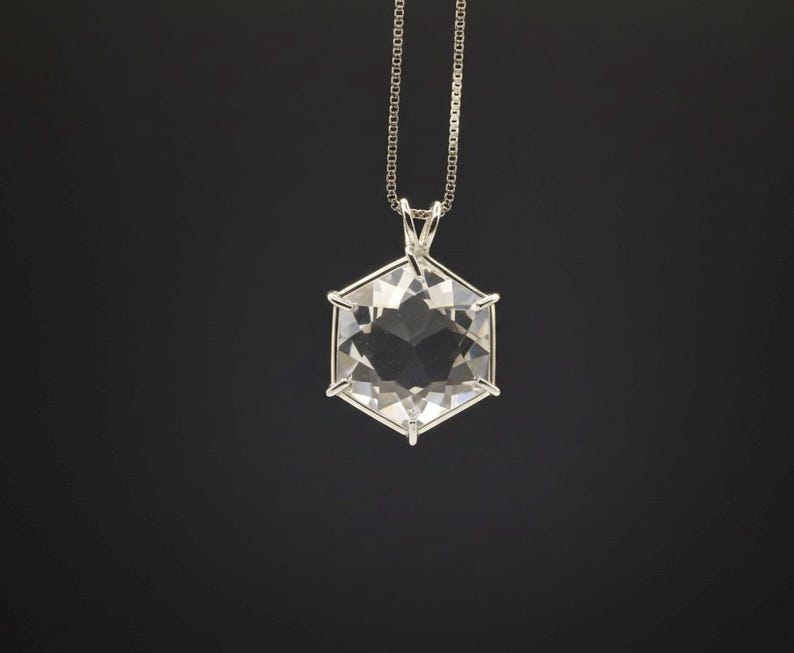 Op de afbeelding: Een heldere, facetgeslepen kristallen hanger in een zeshoekige vorm, hangend aan een fijne zilveren ketting. De hanger is gezet in een zilveren frame, en het kristal reflecteert licht, waardoor een sprankelend effect ontstaat. De achtergrond is effen zwart.