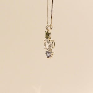May include: A gold-colored necklace with a pendant featuring three gemstones: a green, a clear, and a purple stone. The pendant is suspended from a delicate chain. The background is a solid, light beige color.