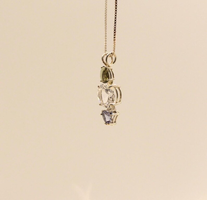 May include: A gold-colored necklace with a pendant featuring three gemstones: a green, a clear, and a purple stone. The pendant is suspended from a delicate chain. The background is a solid, light beige color.