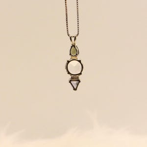 May include: A silver pendant necklace with a geometric design. The pendant features a green teardrop-shaped stone, a large round white stone, and a small triangular white stone. The necklace chain is a delicate silver chain.