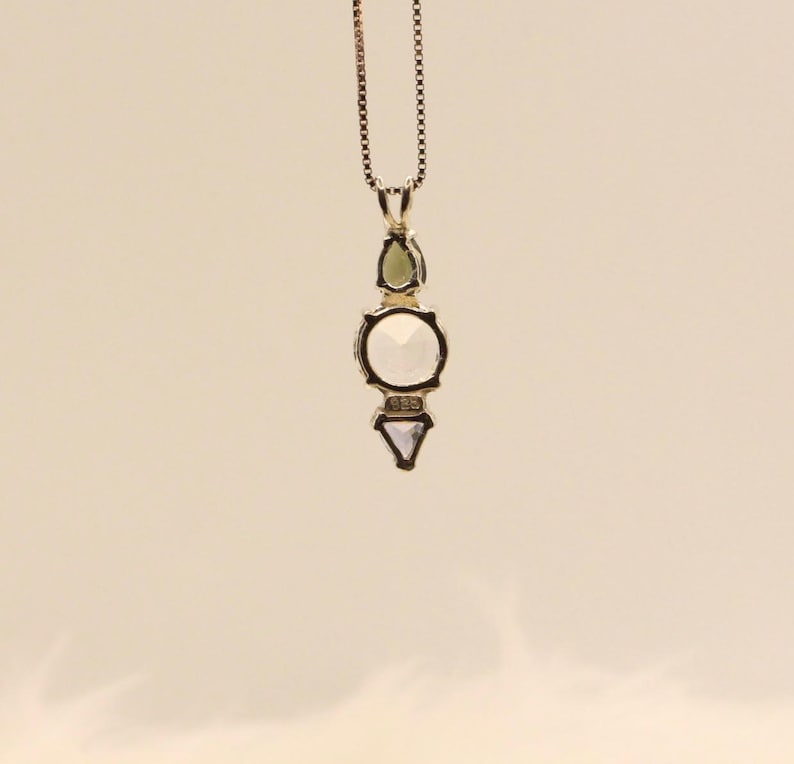May include: A silver pendant necklace with a geometric design. The pendant features a green teardrop-shaped stone, a large round white stone, and a small triangular white stone. The necklace chain is a delicate silver chain.