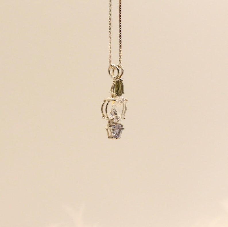 May include: A silver pendant necklace featuring a vertical arrangement of gemstones. The necklace has a delicate chain and showcases a green, clear, and purple gemstone design. The gemstones are faceted and set in a silver metal setting.