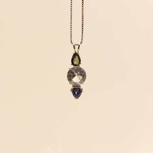 May include: A pendant necklace featuring a silver chain and a vertical arrangement of gemstones. The gemstones include a teardrop-shaped green stone, a large round clear stone, and a small, triangular blue stone. The necklace is displayed against a neutral background.