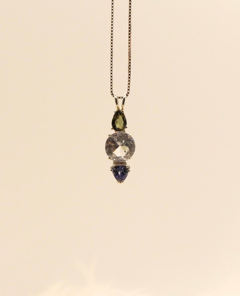 May include: A pendant necklace featuring a silver chain and a vertical arrangement of gemstones. The gemstones include a teardrop-shaped green stone, a large round clear stone, and a small, triangular blue stone. The necklace is displayed against a neutral background.