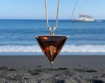 May include: A silver necklace with a large, triangular, brown gemstone pendant. The pendant is suspended from a delicate chain. The background features a beach with black sand, the ocean, and a sailboat.
