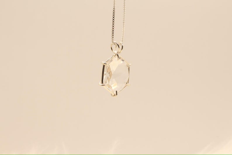 Op de afbeelding: Een zilveren ketting met een heldere, facetgeslepen kristallen hanger. De hanger heeft een geometrische vorm met meerdere zijden en hangt aan een fijne ketting. Het kristal reflecteert het licht, waardoor een sprankelend effect ontstaat. De achtergrond is effen en neutraal.