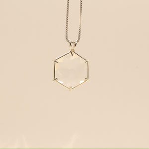 Op de afbeelding: Een zilveren ketting met een geometrische, zeshoekige hanger. De hanger is helder en facetgeslepen, waardoor het licht wordt opgevangen. De ketting is delicaat en hangt aan de bovenkant van de hanger. De achtergrond is effen en neutraal.