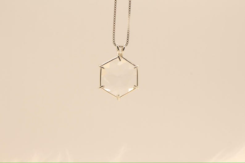 Op de afbeelding: Een zilveren ketting met een geometrische, zeshoekige hanger. De hanger is helder en facetgeslepen, waardoor het licht wordt opgevangen. De ketting is delicaat en hangt aan de bovenkant van de hanger. De achtergrond is effen en neutraal.