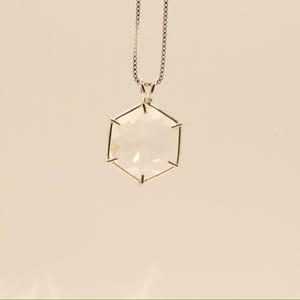 Op de afbeelding: Een zilveren ketting met een heldere, zeshoekige hanger. De hanger hangt aan een fijne ketting. Het geometrische ontwerp en het heldere kristal cre&euml;ren een moderne en elegante uitstraling. De ketting is geschikt voor dagelijks gebruik of speciale gelegenheden.